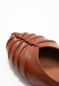 Brązowe skórzane płaskie buty z plisowanym wzorem i plecionym akcentem przy otwarciu. Gładka tekstura w zaokrąglonym kształcie.