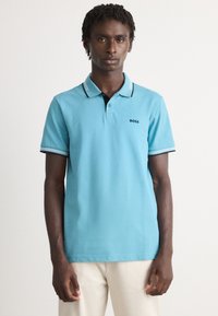 Ανοιχτό μπλε polo φανελάκι από βαμβάκι, με μαύρο γιακά, πλαισιωμένο από τρία κουμπιά και ριγέ λεπτομέρειες στα μανίκια. Εμφανές το λογότυπο "BOSS".