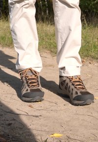 Buty trekkingowe z szarym siateczkowym wierzchem, wzmocnionym czubkiem, kontrastującymi brązowymi sznurowadłami i czarną gumową podeszwą, ustawione na ścieżce żwirowej.