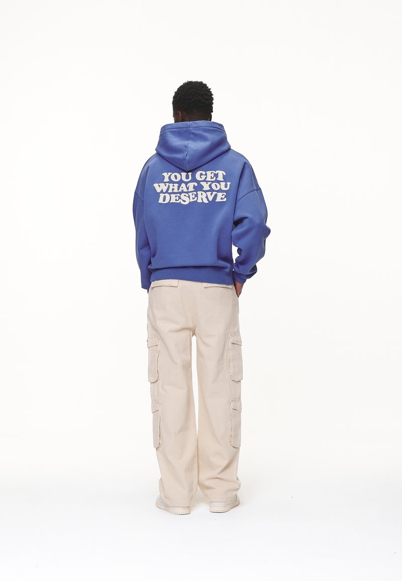Sudadera azul con texto blanco que dice "TÚ OBTIENES LO QUE MERECES" en la parte de atrás, combinada con pantalones cargo beiges y zapatillas blancas.