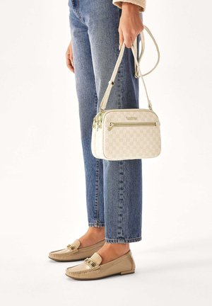 Sac bandoulière blanc à motifs avec deux compartiments zippés, en matériau texturé, et une sangle légère, associé à un jean bleu et des mocassins beiges.