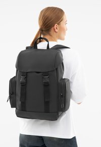 Svart ryggsäck gjord av glatt material, med lockstängning, två sidofickor och justerbara axelremmar. Enkel, funktionell design.