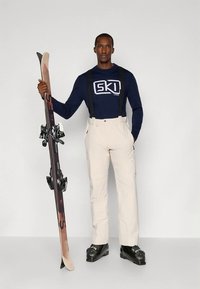 Hiihtoasu, jossa on laivastonsininen pitkähiha paita, jossa "SKI"-grafiikka, beiget haalarihousut henkselin kanssa, mustat hiihtokengät ja puiset sukset.