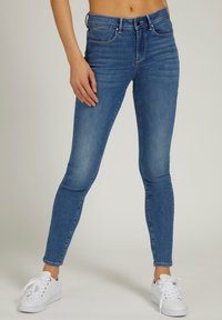 Vaqueros de denim azul skinny-fit con cinco bolsillos, cierre de botón y ligero desvanecimiento. Combinados con zapatillas blancas.