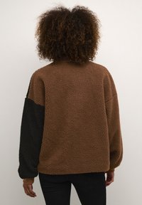 Kiharatukkainen henkilö, joka pukeutunut ruskeaan fleece-takkiin, jossa on yksi musta hiha, seisoo yksivärisen vaalean taustan edessä.