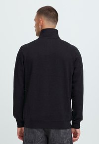 Uomo con capelli corti e orecchino ad anello d'argento, indossante un felpa nera a collo alto, visto di spalle su uno sfondo chiaro e semplice.