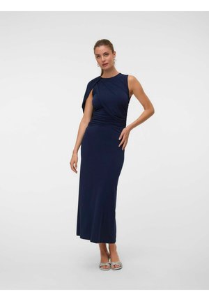 Femme posant avec une main sur la hanche, portant une robe maxi sans manches bleu marine avec un détail drapé sur une épaule et des sandales blanches.