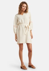 Crème kleurige jurk met een V-hals, lange mouwen en een strikceintuur in de taille. Bevat geborduurde cirkelvormige accenten op de mouwen. Gladde textuur.