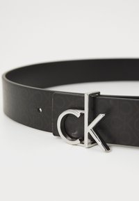 Černý kožený pásek s vyraženým vzorem loga a stříbrnou kovovou sponou s designem "CK".