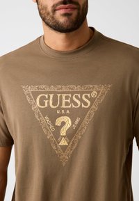 Brązowa bawełniana koszulka z złotym trójkątnym logo z napisem "GUESS U.S.A. WASHED JEANS" oraz ozdobnymi akcentami wokół.