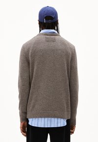 Brauner strukturierter Pullover mit langen Ärmeln, einem runden Halsausschnitt und einem kleinen Logo auf dem Rücken, über einem hellblau gestreiften Hemd getragen.