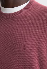Burgundy knitted sweater with a textured finish, featuring a white crew neck layer underneath and a subtle embossed logo near the hem.