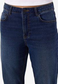 Tamsiai mėlyni džinsai iš džinso medžiagos su klasikiniu kirpimu, priekyje užsegami sagomis, turi penkias kišenes ir kontrastingas siūlių detales.