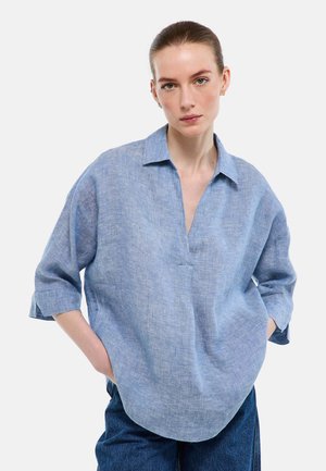Femme aux cheveux attachés portant une chemise ample en lin bleu clair à manches 3/4 et un jean bleu foncé, les mains dans les poches, fond neutre.