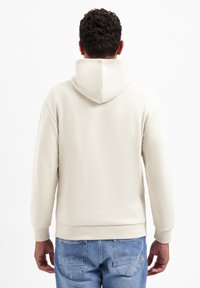 Crèmekleurige hoodie met een gestructureerd ribbelpatroon, voorzien van een trekkoordkap en verharde mouwen, gedragen met blauwe jeans.