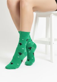 Piernas con calcetines verde brillante con patrones de cactus negros, descansando cerca de un taburete de madera blanca sobre un suelo blanco y liso.