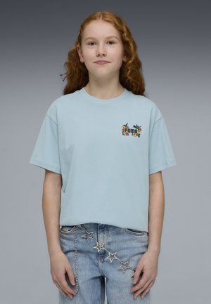 Joven con largo cabello rizado rojo, lleva una camiseta azul claro de Puma y unos vaqueros azul claro con un cinturón de cadena con estrellas, sobre un fondo gris.