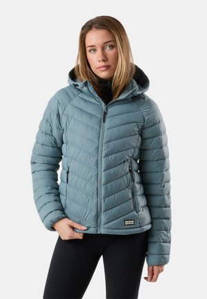 Młoda kobieta w jasnoniebieskiej pikowanej kurtce z kapturem i przednim zamkiem błyskawicznym oraz czarnych spodniach, stojąca na białym tle.