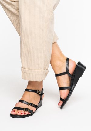 Black leather sandals with multiple straps, featuring gold-tone buckle accents and a low wedge heel. Worn with beige corduroy pants.