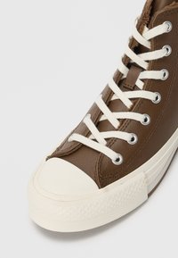 Barna magas szárú sneaker fehér gumitalppal és orral, fehér fűzővel, fémből készült fűzőlyukakkal, valamint látható barna bélésű bolyhos béléssel.
