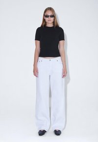 Top negro ajustado de manga corta combinado con unos pantalones anchos blancos de pierna ancha, complementado con unas pequeñas gafas de sol negras y zapatos oscuros.