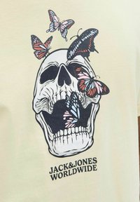 Korte mouw lichtgele t-shirt met een grafisch ontwerp van een schedel met kleurrijke vlinders. Het ontwerp bevat zwart en roze accenten.