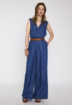 Femme aux cheveux châtain clair portant une combinaison en denim bleu sans manches, ceinture marron, bracelets et bagues en or, debout devant un fond clair et uni.