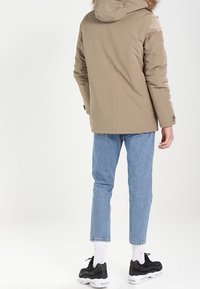 Giacca invernale beige con cappuccio e rifinitura in pelliccia, realizzata in un materiale liscio e resistente all'acqua. Abbinata a jeans blu chiaro e sneakers nere.