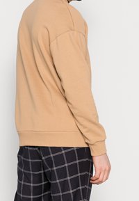 Béžová mikina s dlouhými rukávy a žebrovanými manžetami, s volným střihem a měkkou texturou. V kombinaci s černými kostkovanými kalhotami.