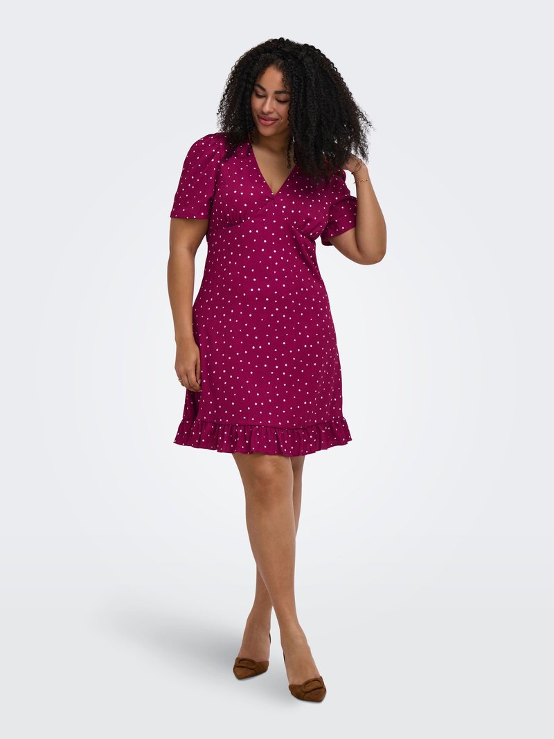 Vrouw met krullend haar die een paarse polkadotjurk met ruches aan de zoom draagt en bruine puntige schoenen, staand en haar haar aanrakend.