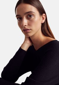 Zbliżenie osoby o gładkiej skórze, ubraną w czarny sweter prążkowany z dekoltem w serek, opierającą podbródek na dłoni. Tło jest białe.