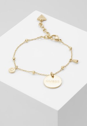Delicaat gouden kettingarmband met kleine kralen, een ovale tag gegraveerd met "GUESS", een ronde tag met een "G" en een sluiting met karabijnhaak.