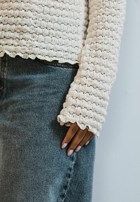 Lichtbeige gebreide top met textuur en gerimpelde randen, gecombineerd met donkerblauwe denim. Close-up van de mouw en hand die op de stof rust.