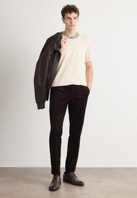 Un t-shirt beige à col rond, une veste bomber noire, un pantalon en velours côtelé sombre et des chaussures à lacets brunes, se tenant sur un sol en béton devant un fond clair.