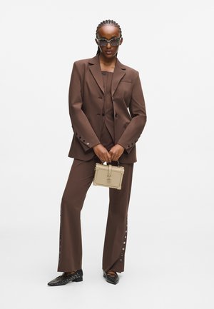 Completo in marrone con giacca su misura e dettagli sui bottoni. Abbinato a una borsa beige e a ballerine nere con borchie. Gli occhiali da sole neri completano il look.