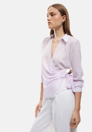 Giovane donna con una camicetta a portafoglio viola chiaro testurizzata con un grande bottone dorato, abbinata a pantaloni bianchi a vita alta, in piedi di profilo.