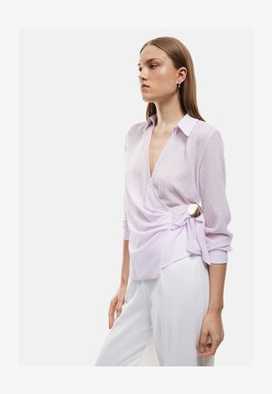 Giovane donna con una camicetta a portafoglio viola chiaro testurizzata con un grande bottone dorato, abbinata a pantaloni bianchi a vita alta, in piedi di profilo.