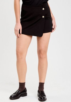 Kvinde iført en sort asymmetrisk mini nederdel med to dekorative knapper, brune sokker og sorte loafers mod en ensfarvet baggrund.