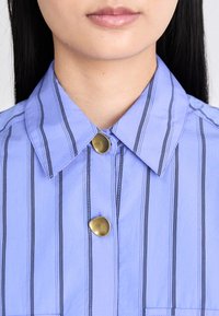 Collared shirt in light blue with black vertical stripes, featuring a brass button at the collar and a chest pocket. Smooth texture.