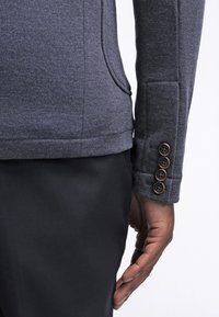 Mörkgrå tygjacka med en slät yta, med en sidoficka och en fyraknappar detalj vid manschetten. Bärs över svarta byxor.