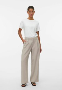 Ženska stoji z eno roko v žepu, oblečena v belo kratko srajco, bež široke hlače in črne sandale z odprtim prstom na belem ozadju.