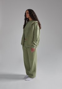 Set da loungewear a due pezzi di colore verde oliva, composto da una felpa con zip a vestibilità comoda e pantaloni ampi coordinati, realizzati in tessuto morbido.
