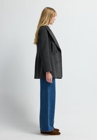 Abrigo gris de gran tamaño con doble botonadura y un diseño estructurado, exhibido junto a unos vaqueros de pierna recta azul y calzado marrón.