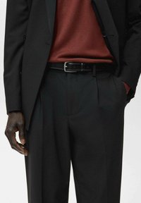 Mężczyzna w czarnych spodniach i marynarce, z brązowym swetrem włożonym w spodnie, czarnym paskiem, z jedną ręką w kieszeni, a drugą swobodnie opadającą wzdłuż ciała.