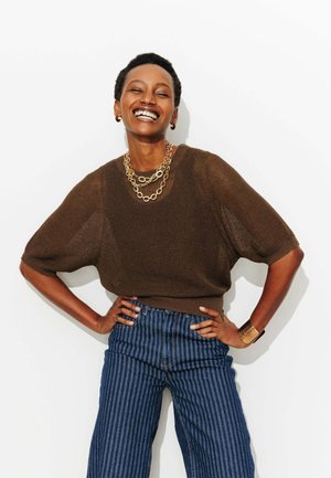 Personne souriante aux cheveux courts portant un haut en tricot marron, des colliers en chaîne dorée, un pantalon large à rayures bleues, et un bracelet manchette en or, mains sur les hanches.