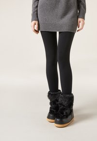 Schwarze Stiefel aus Kunstfell mit einer strukturierten Oberseite, Gummisohle und cremefarbener Außensohle. Kombiniert mit schwarzen Leggings und einem grauen Pullover.