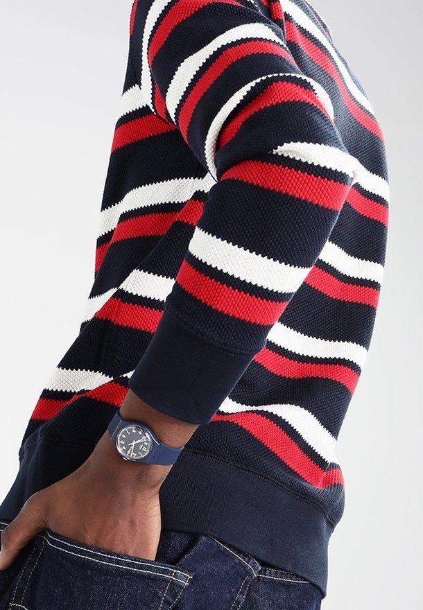 Personne portant un pull à rayures bleu marine, rouge et blanc avec une montre bleue au poignet, main dans la poche d'un jean en denim foncé, sur un fond uni.