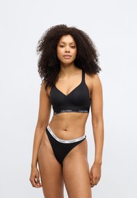 Crni pamučni bralette s V-izrezom i tankim naramenicama, s brendiranim elastičnim pojasom. Usklađeni crni tanga gaćice s bijelim logotipom na pojasu.