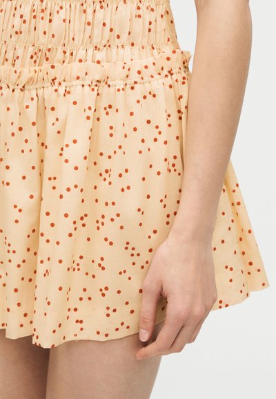 Μπεζ φούστα με κόκκινα πουά, με λάστιχο στη μέση που δημιουργεί πιέτες, φορεμένη από άτομο με ορατό χέρι και παλάμη που ακουμπούν κοντά στο τελείωμα της φούστας.