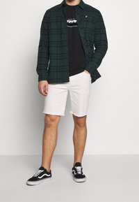 Zeleno-modrý kostkovaný košile na knoflíky přes černé tričko, spárovaná s bílými denimovými šortkami a černými teniskami s bílými detaily.